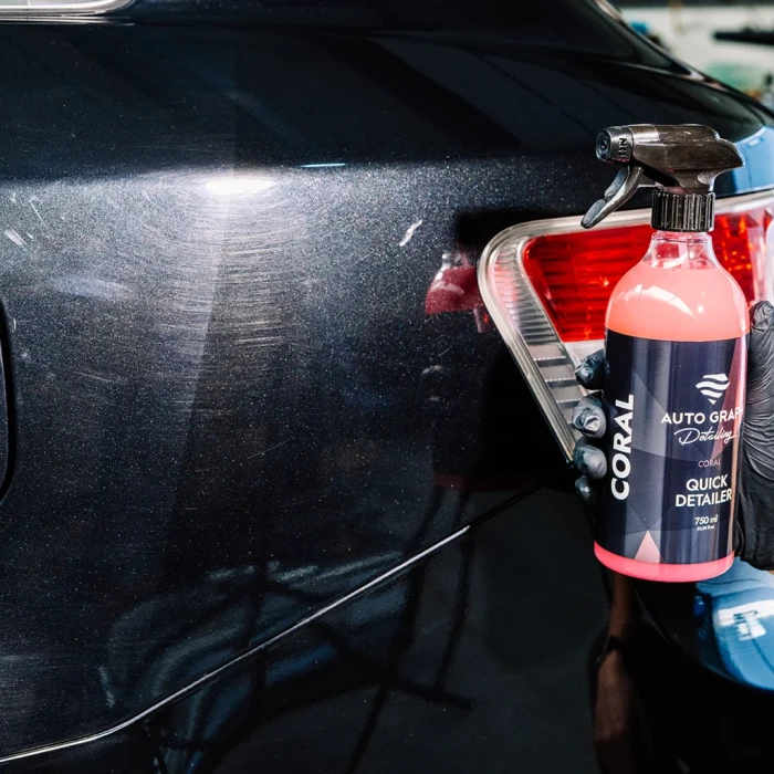 Ręka trzyma butelkę CORAL Quick Detailer przy tylnej lampie ciemnego samochodu