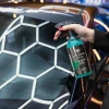 dłoń trzyma butelkę EMERALD Hydrophobic Glass Cleaner przy szybie oklejonej folią w plaster miodu